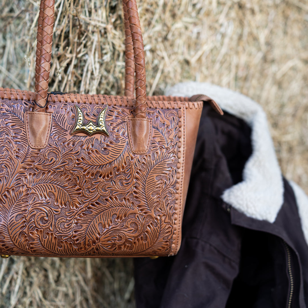 A brown tooled leather handbag with braided shoulder straps and a gold-tone horn emblem, hanging against a hay bale background next to a black jacket with a white shearling lining.