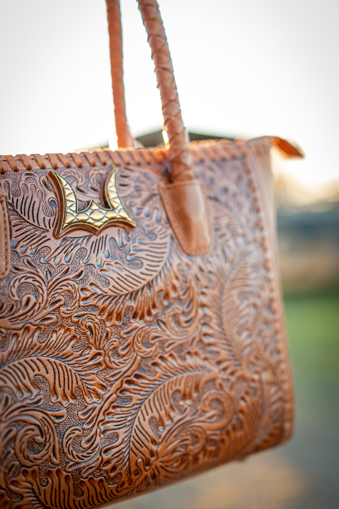 A close-up of a brown tooled leather tote bag with intricate floral embossing, a gold-toned bat-shaped hardware accent, and braided shoulder straps, photographed outdoors against a softly blurred background.