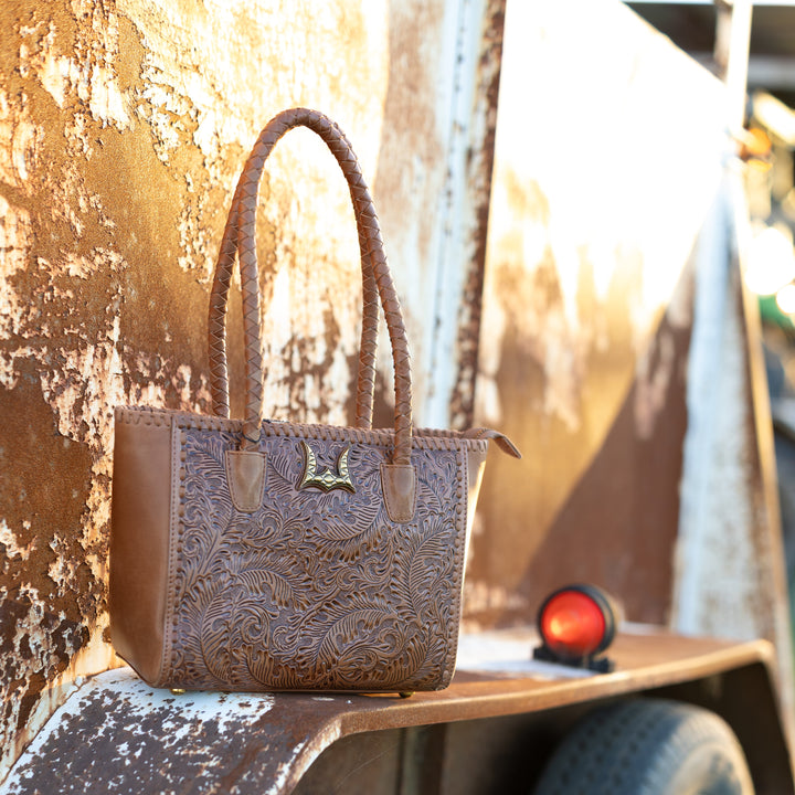 A brown tooled leather handbag with double handles sits on the rusty bed of a vintage truck trailer next to a tire.