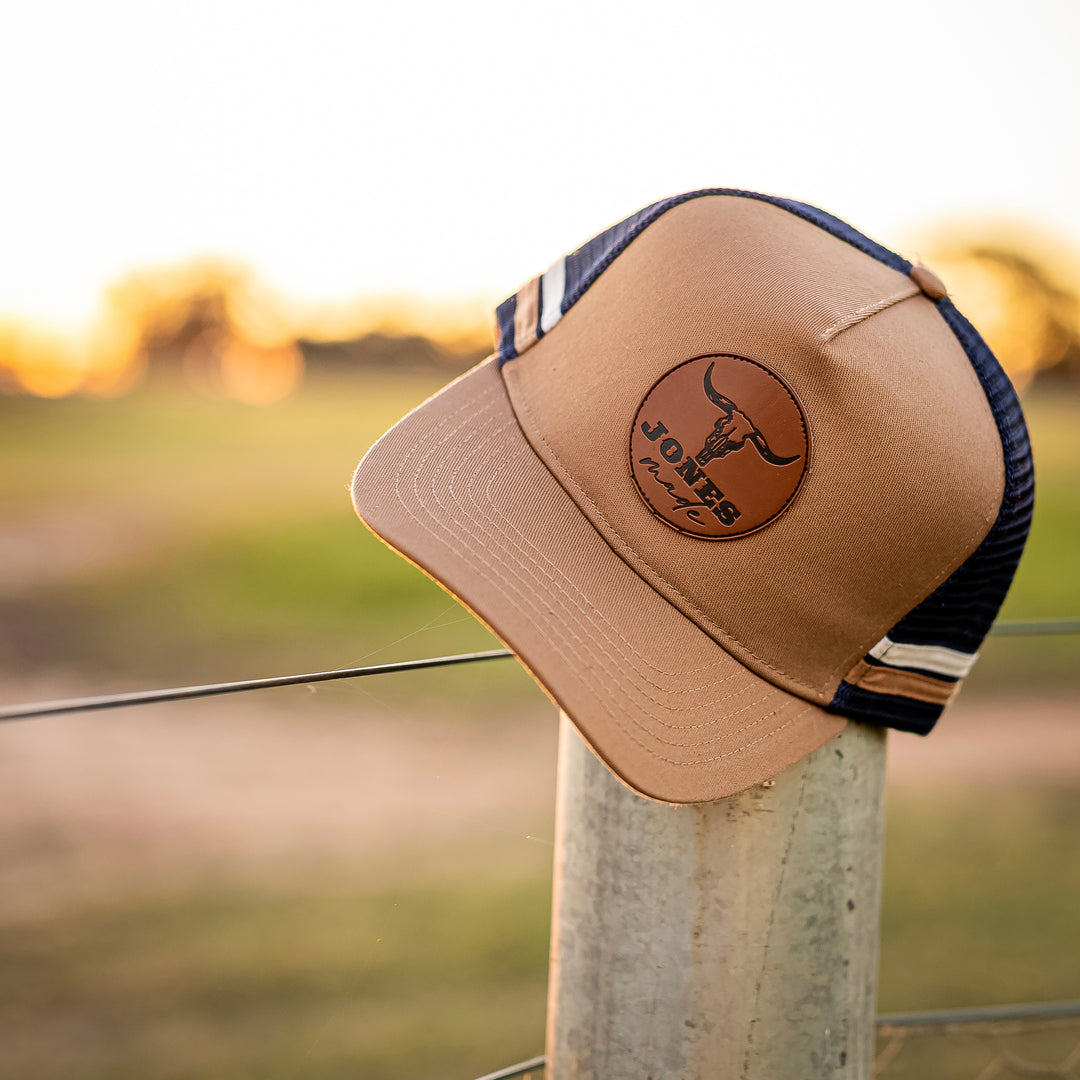 A tan and navy blue trucker hat with a circular brown leather patch on the front, resting on a metal fence post outdoors at sunset.