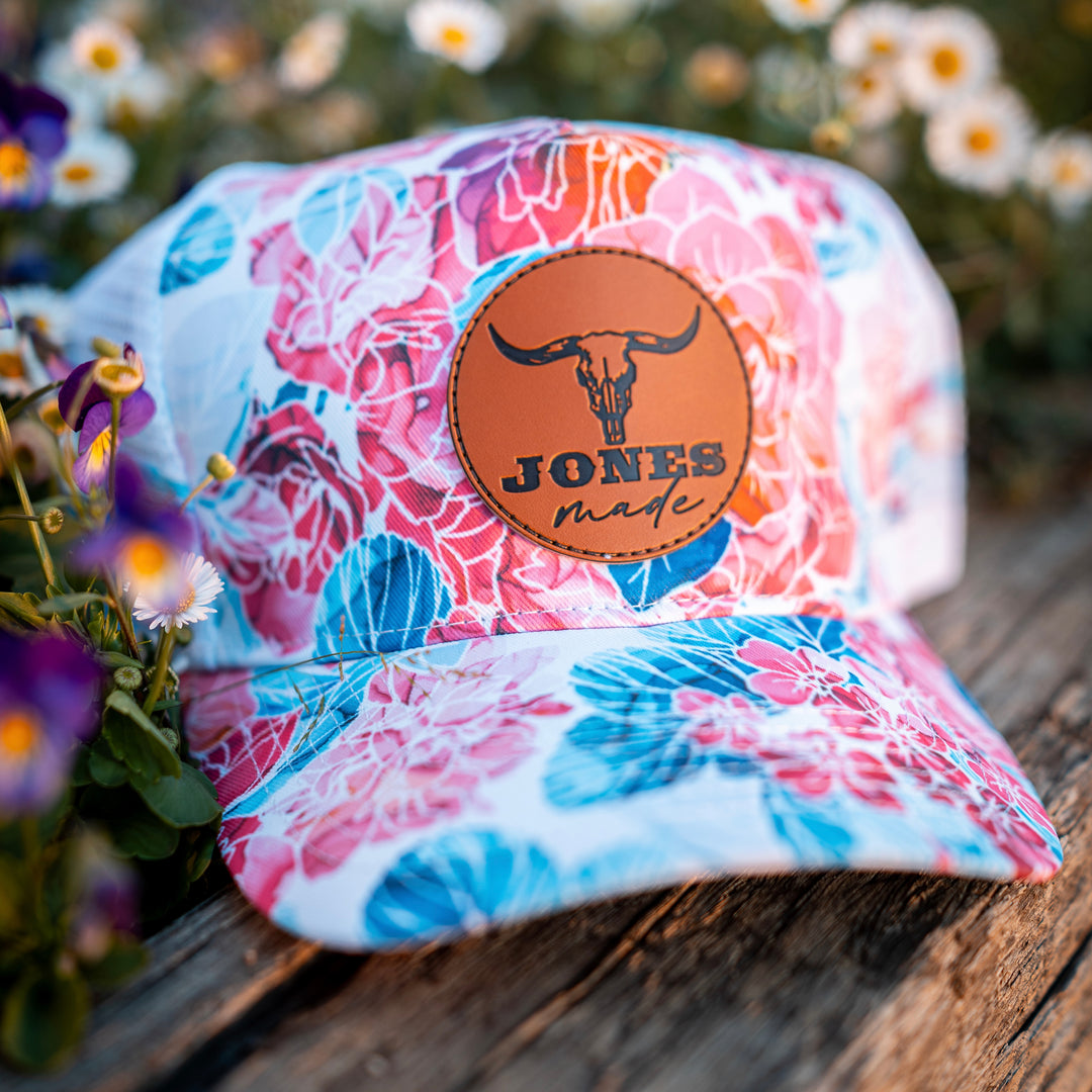 A multicolor floral trucker cap with a leather  'JONES made.' patch on the front, resting on a rustic wooden surface with blurred flowers in the background.