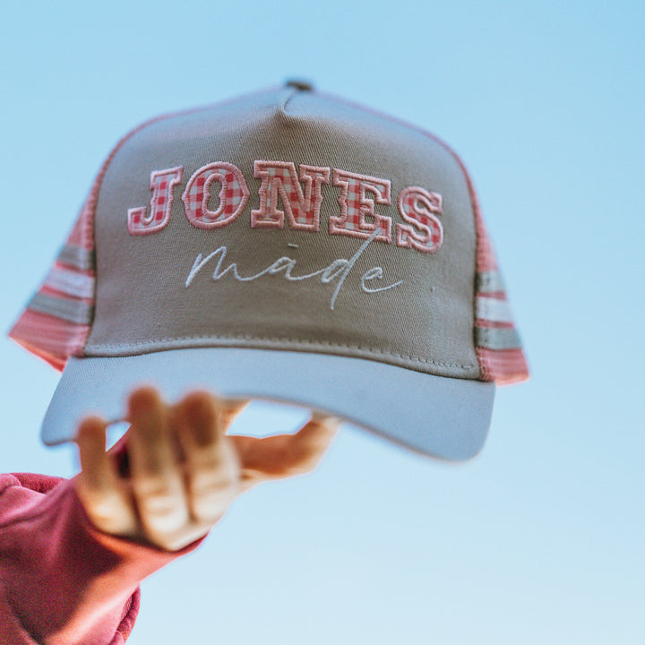 A person's hand holds up a gray trucker hat with pink and white gingham lettering that reads 'JONES made' against a clear blue sky.