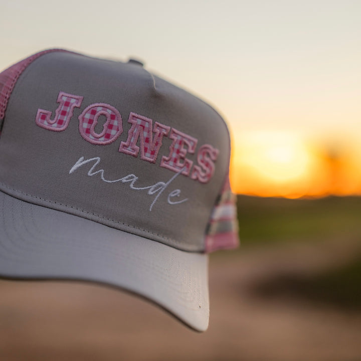 A gray and pink gingham trucker hat with 'JONES made' embroidered on the front, set against a blurred sunset background.