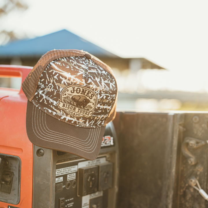 A brown and beige camouflage trucker hat with a 'Jones bison tours' logo on the front, resting on top of a portable generator in the bed of a pickup truck.