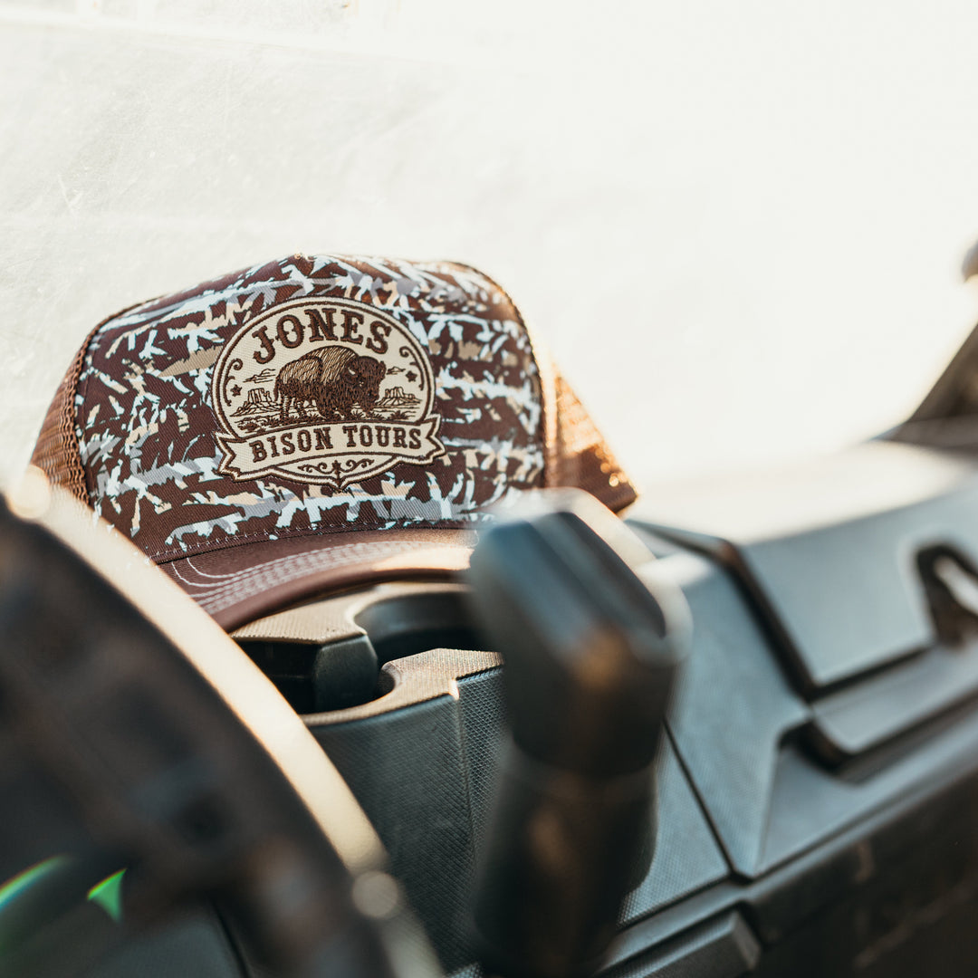 A brown and beige patterned trucker hat with a circular 'Jones Bison Tours' logo patch, resting on the dashboard of a vehicle.