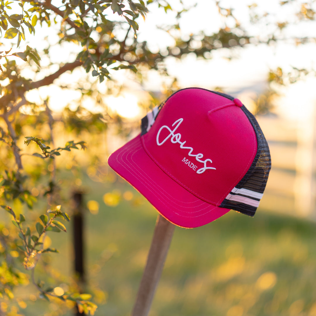 A bright pink trucker hat with white script embroidery reading 'Jones madel' hanging from a wooden post outdoors. The hat has a pink front panel, white script logo, and navy mesh back with white and pink stripes.