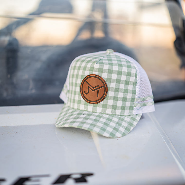 A green and white gingham trucker hat with a brown circular leather patch on the front, resting on the hood of a white vehicle.