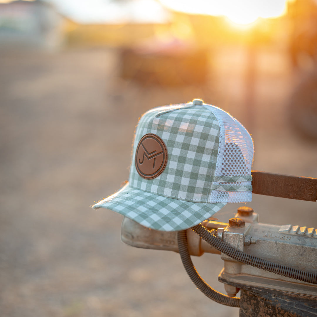 A light green and white gingham trucker hat with a brown leather circular patch on the front, resting on the edge of a rusty metal hitch in an outdoor farm setting at sunset.