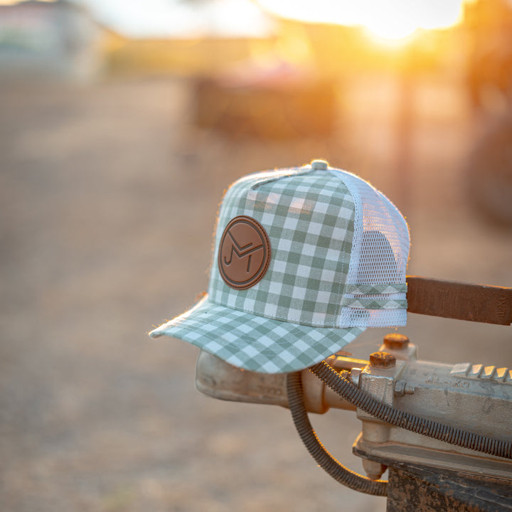 A light green and white gingham trucker hat with a brown leather circular patch on the front, resting on the edge of a rusty metal hitch in an outdoor farm setting at sunset.