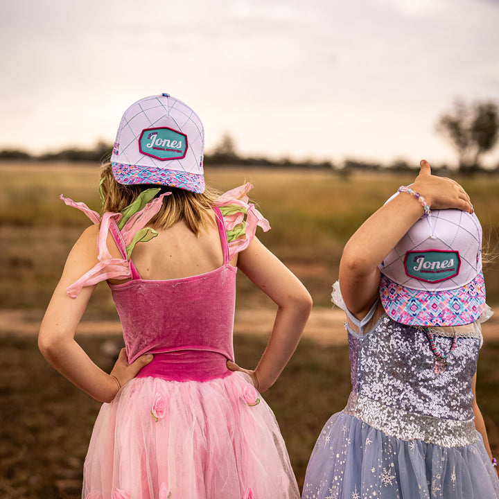 Two children seen from behind wearing matching light purple baseball caps with 'Jones Made' on the back, along with colorful dresses in pink and blue, standing outdoors in a grassy field under a cloudy sky.