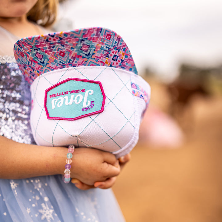 A child in a light blue tulle dress with white snowflake patterns holds a cap. The cap has a white diamond-quilted pattern, a pink stitched border, and a turquoise center patch with the name 'Jones made' in script.