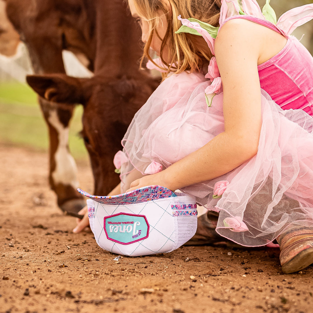 A young girl in a pink fairy costume kneels on a dirt path next to a brown cow, filling a cap with rocks. The background is a soft-focus outdoor setting with green foliage.