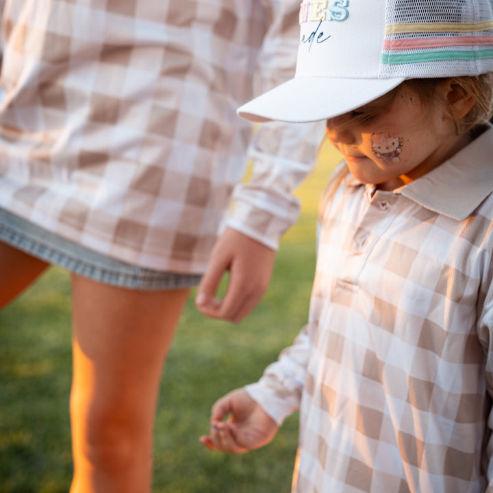A child wearing a white trucker hat with pastel stripes and a matching beige-and-white gingham long-sleeve shirt, standing outdoors on grass at sunset while an adult in a matching gingham top stands beside them.