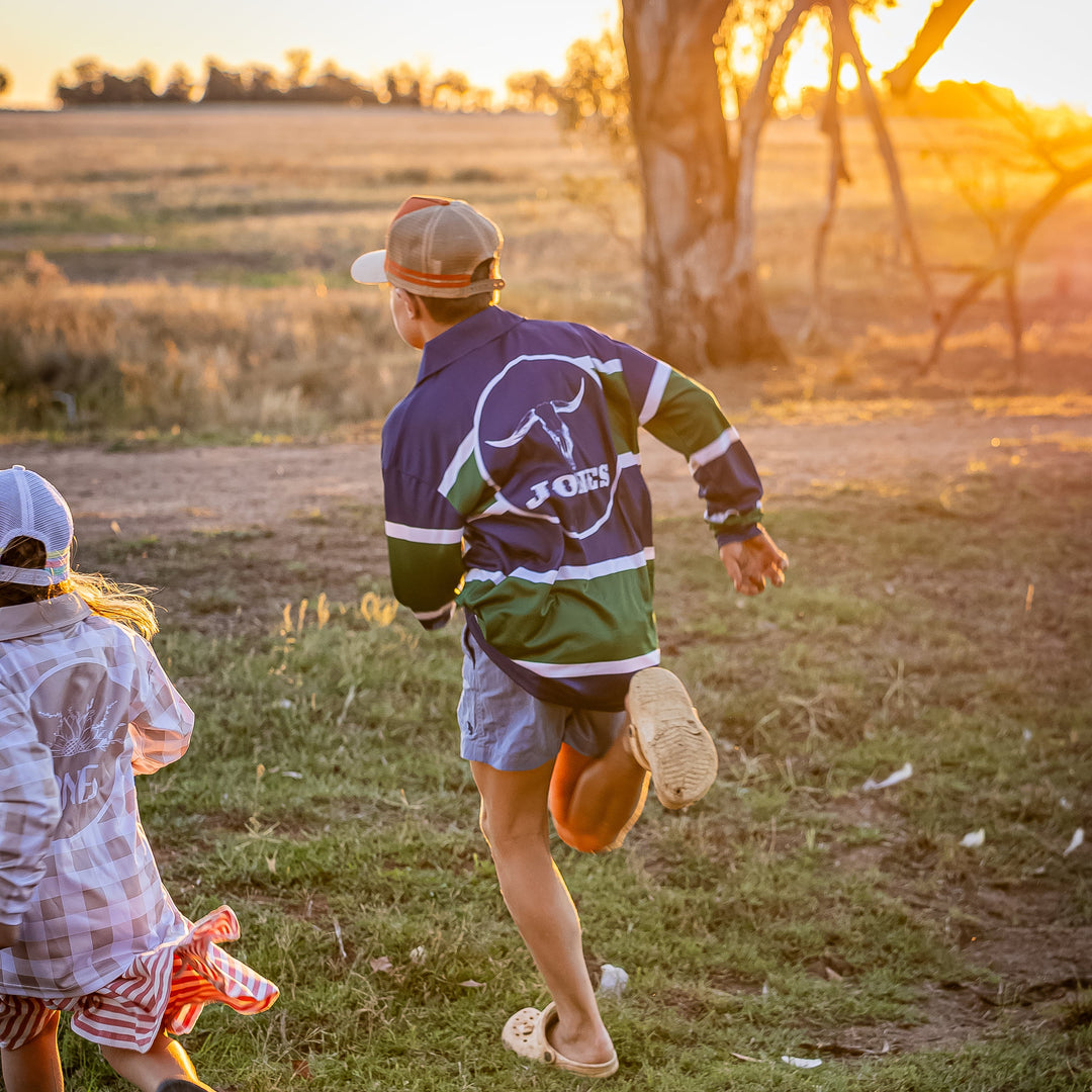 A child wearing a dark navy and green long-sleeve rugby jersey with a white buffalo logo and 'JONES' on the back, running outdoors in a grassy field at sunset. Another child is partially visible on the left.