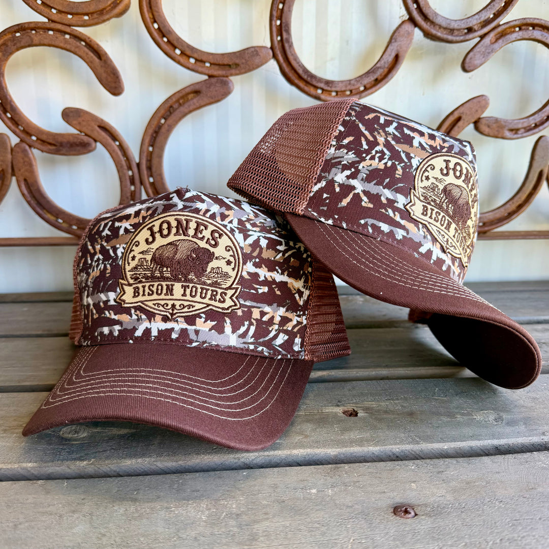 Two brown and beige camouflage trucker hats with a circular 'Jones Bison Tours' logo on the front, displayed on a rustic wooden surface with a decorative metal horseshoe wall in the background.