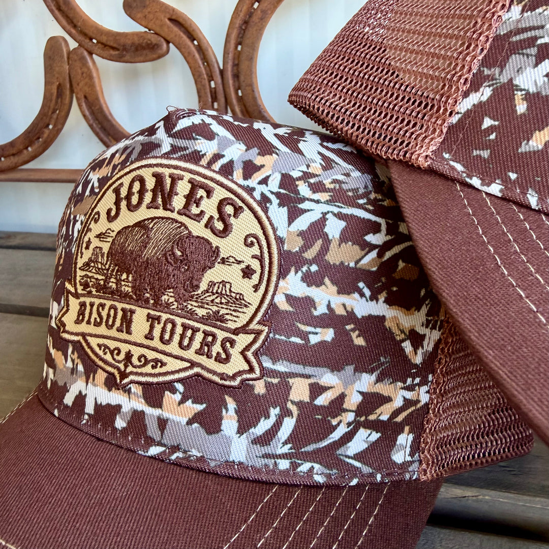 A brown and beige camouflage trucker hat with a brown embroidered patch reading 'JONES BISON TOURS' and a buffalo graphic, shown alongside another matching hat on a rustic wooden surface.