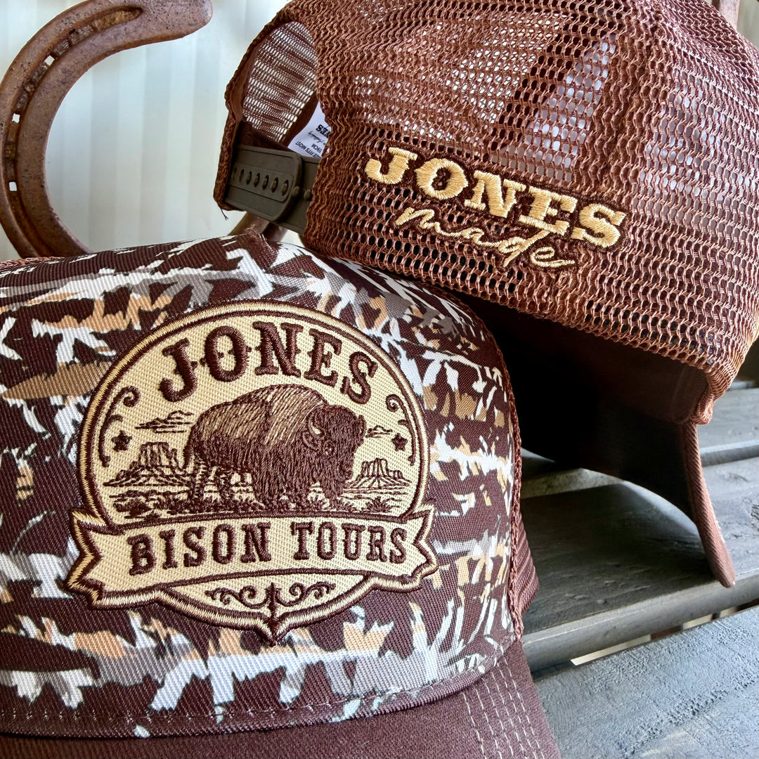 Two branded trucker-style caps displayed together: one in brown mesh with gold embroidered 'JONES MADE' text, and one in brown, beige, and white camouflage with a large 'JONES BISON TOURS' patch on the front. 