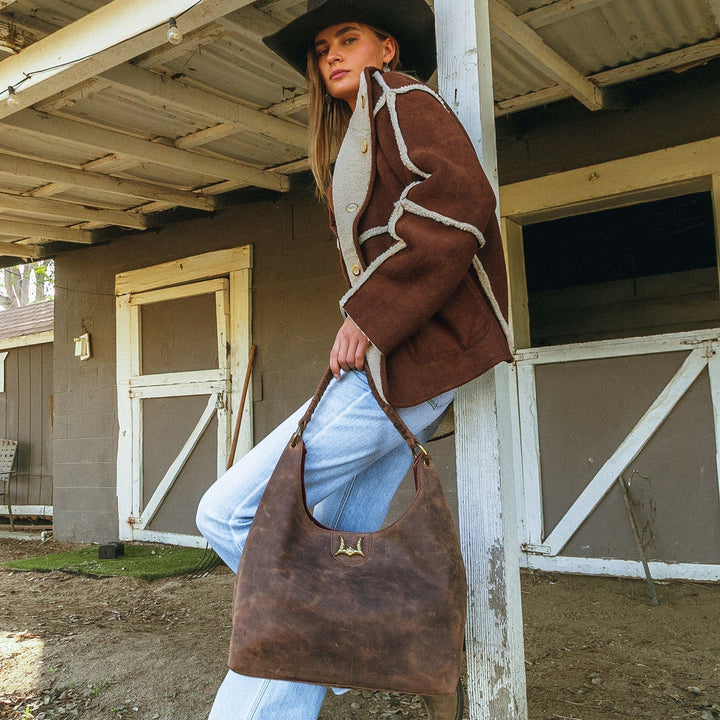 Person wearing a brown jacket and hat, holding a leather bag in front of a wooden building.