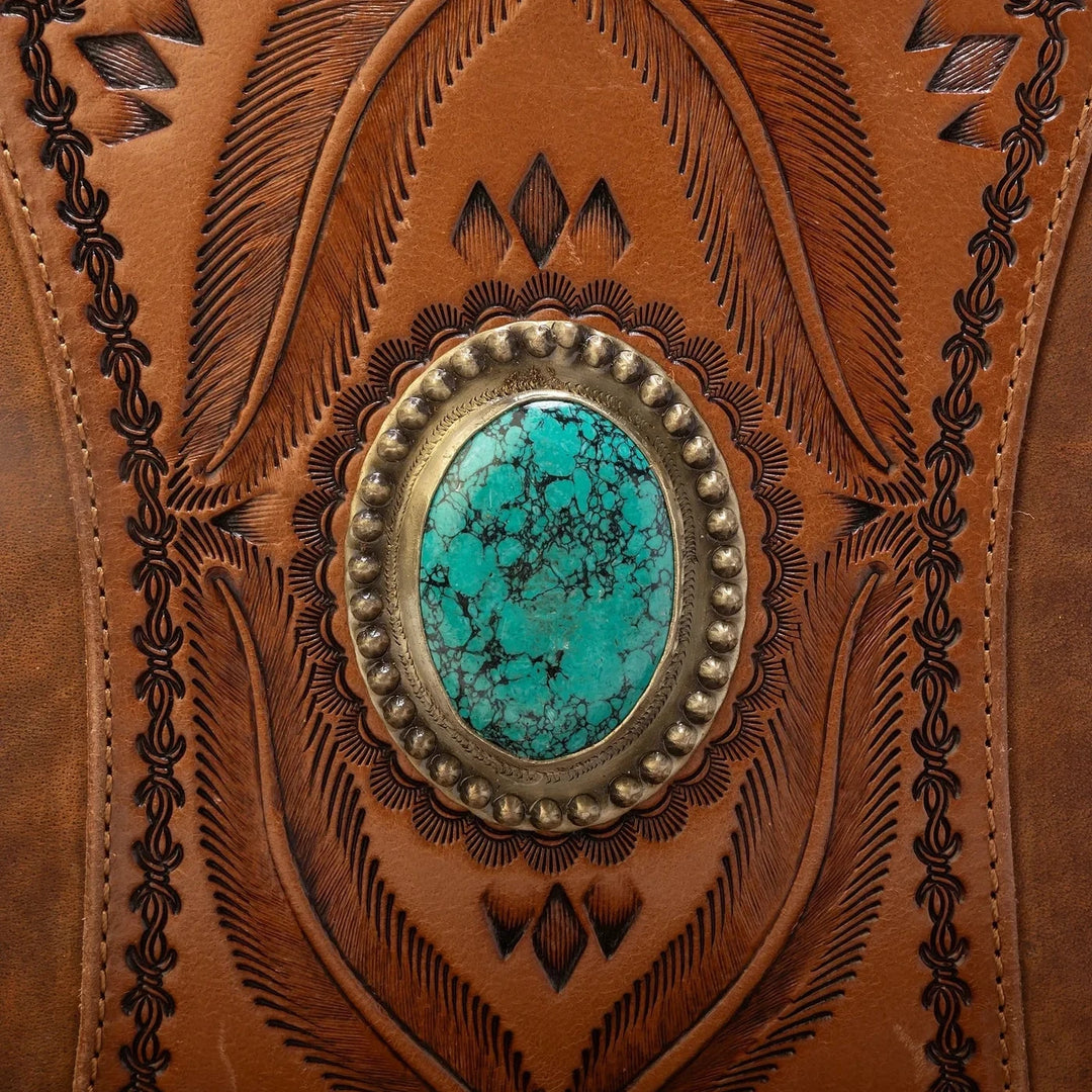 Close-up of a brown tooled leather item featuring an oval turquoise stone set in a silver-toned metal bezel, surrounded by decorative embossed patterns.
