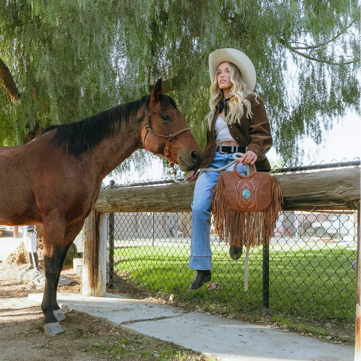 Woman in cowboy hat and boots standing next to a horse, holding a brown leather bag with fringe.