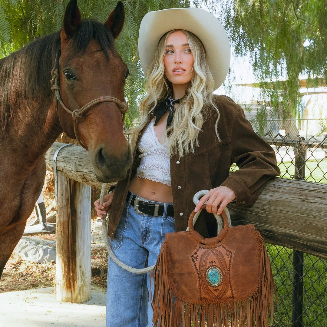A woman in western attire, including a white cowboy hat, brown jacket, and blue jeans, stands next to a brown horse while holding a brown leather fringe handbag with a turquoise accent. The setting is outdoors under a tree with a fence in the background.