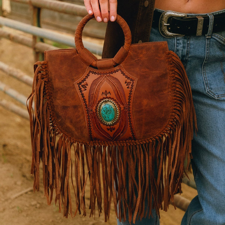 Person holding a brown leather handbag with fringe and a turquoise stone, wearing jeans and a jacket.