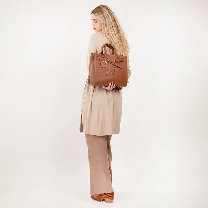 A woman with long blonde hair is shown from the back, wearing a beige trench coat and wide-leg trousers while carrying a brown leather tote bag over her shoulder, set against a plain white studio background.