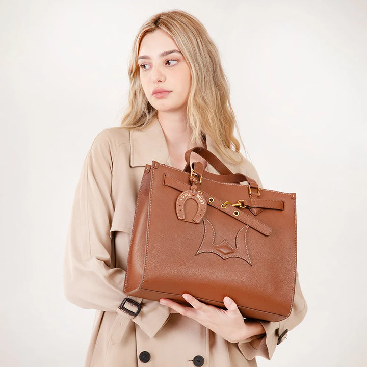 A woman in a beige trench coat holds a brown leather tote bag with a decorative horse-head charm and gold-tone hardware against a plain light background.