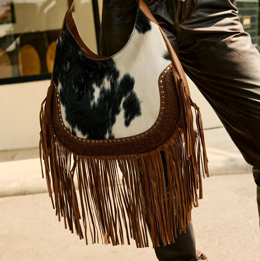 Person holding a cow print handbag with fringe details outdoors.