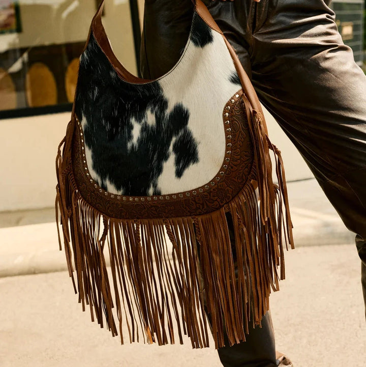 Person holding a cow print handbag with fringe details outdoors.