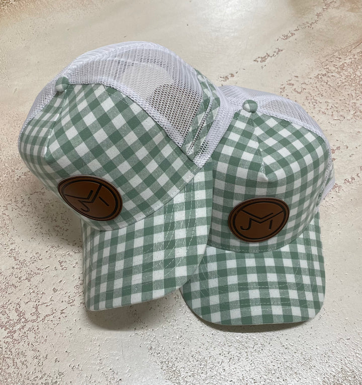 Two green and white gingham trucker caps with a leather logo on the front, available in high profile style.