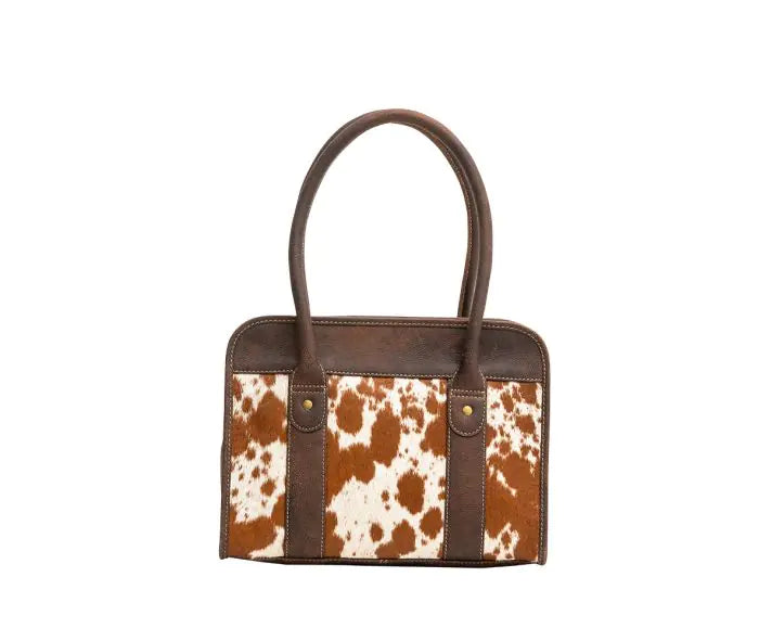 A structured handbag with a brown and white cowhide patterned front panel and dark brown leather handles and trim, shown against a plain white background.