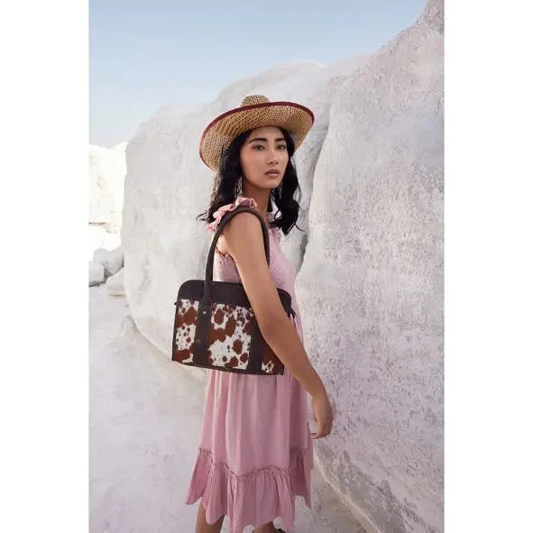 A woman wearing a straw hat and a pink dress stands outdoors against a white rock formation, carrying a brown handbag with a white and brown cow-print pattern.